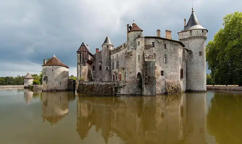 photographie d'un château qui se reflète dans l'eau qui l'encercle. Présence d'un ciel gris menaçant et de quelques arbres à l'arrière plan